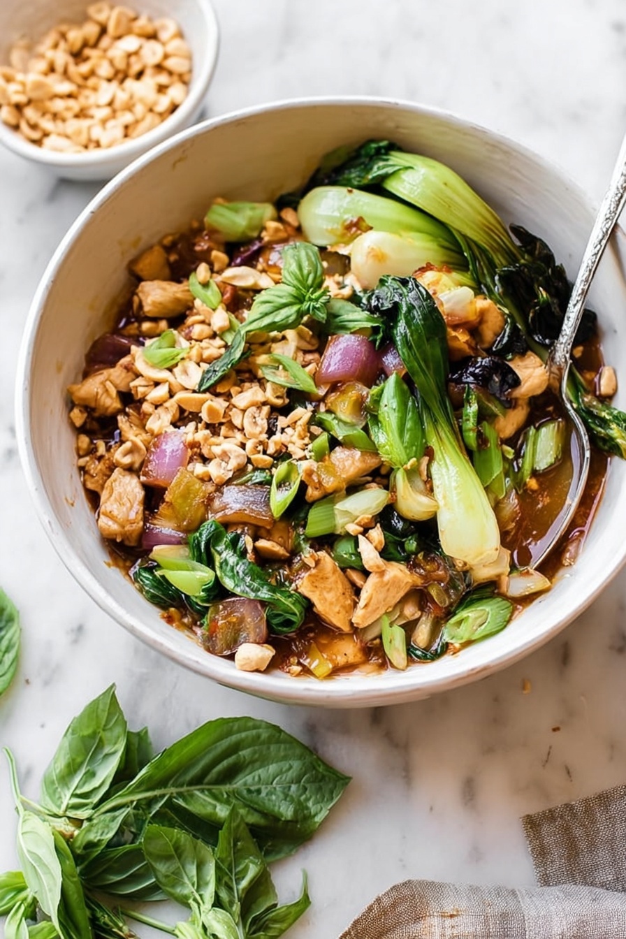 A bowl filled with a colorful stir-fry that has three main layers: the bottom layer contains light brown sauce with pieces of cooked light tan chicken and bright green vegetables like bok choy and fresh basil leaves; the middle layer has red onion slices and dark green leafy bits mixed in; the top layer is sprinkled with crushed light tan peanuts and fresh basil leaves, adding texture and color contrast. The bowl is white ceramic, set on a white marbled surface, with a silver spoon standing inside. Nearby, a small white bowl holds more crushed peanuts, and a few fresh basil leaves lay on the surface. Photo taken with an iphone --ar 2:3 --v 7