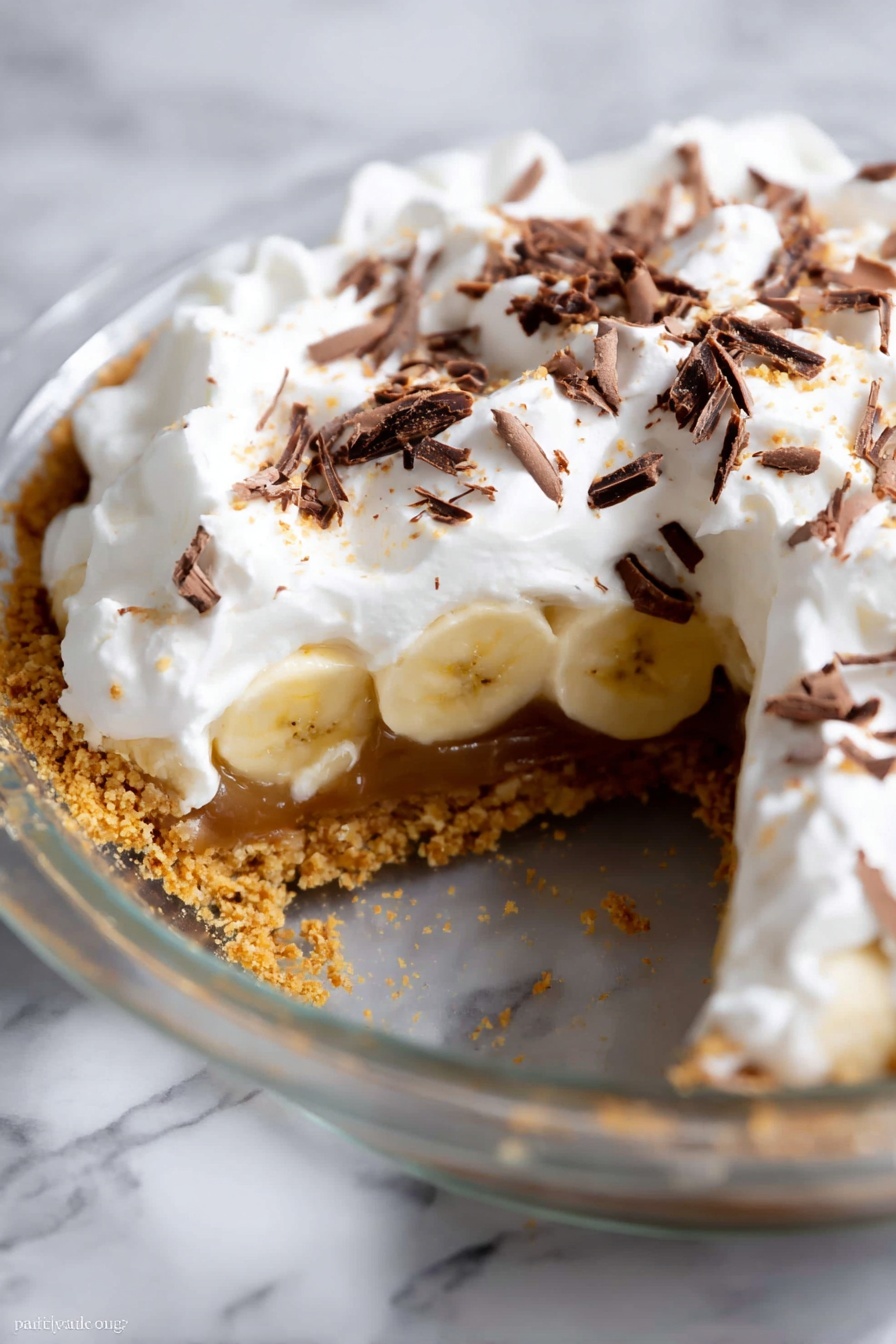 The image shows a close-up of a partially eaten pie in a clear glass pie dish, placed on a white marbled surface. The pie has four visible layers: the bottom layer is a crumbly golden-brown crust, above that a sticky caramel layer, then a layer of sliced bananas arranged in a single row, and finally a thick, fluffy white whipped cream topping with small piles and peaks. On top of the whipped cream, there are thin dark brown chocolate shavings scattered unevenly. The edge of the pie is slightly crumbly where a slice has been removed, revealing the inside layers clearly. photo taken with an iphone --ar 2:3 --v 7
