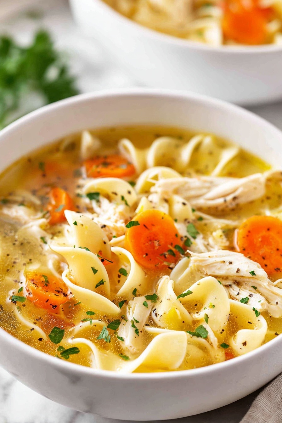 A close-up view of a white bowl filled with chicken noodle soup, held gently by two woman's hands with glittery pink nail polish. The soup has three visible layers: a clear golden broth at the bottom, chunky orange carrot slices and light green celery pieces scattered in the middle, and shredded white chicken with flat, curly egg noodles floating on top, all sprinkled with fresh green parsley and black pepper. The bowl is set against a white marbled background, with a silver spoon seen beside it. photo taken with an iphone --ar 2:3 --v 7