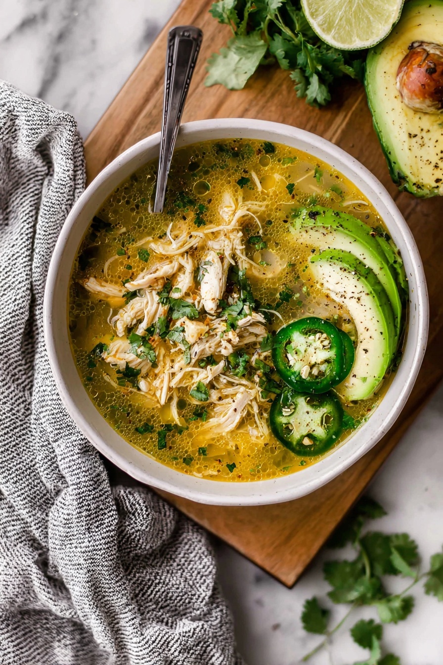 A bowl filled with shredded chicken soup sits on a white marbled surface on a wooden board. The soup is yellowish with small green herbs mixed in, and shredded chicken pieces are visible throughout. Three green jalapeño slices and a fanned avocado slice with black pepper rest on one side of the bowl, next to a halved lime. A silver spoon is inside the bowl, leaning on the edge. There is a gray and white striped cloth under the bowl and some cilantro leaves scattered nearby. Photo taken with an iphone --ar 2:3 --v 7