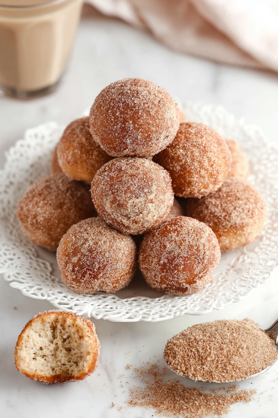 A white plate with detailed lace-like edges holds a stack of round donut holes coated with cinnamon sugar, showing a light brown, slightly rough texture on each piece; one donut hole is in front of the plate, broken open to reveal a soft, fluffy inside with a pale golden color, while a silver spoon filled with cinnamon sugar sits beside the donuts on a white marbled surface, creating a cozy and sweet scene, photo taken with an iphone --ar 2:3 --v 7