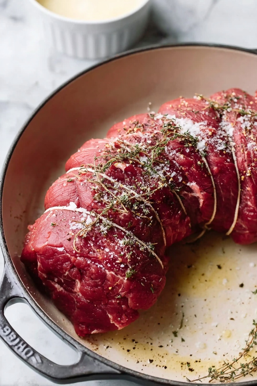 A tied raw meat roast with a curved shape sits in a white cast iron pan. The roast is deep red with some fat showing and is seasoned with a light sprinkling of coarse salt and dried herbs scattered on top. The pan has a sturdy handle, and the background is a white marbled surface with a small white bowl of creamy sauce nearby. Photo taken with an iphone --ar 2:3 --v 7