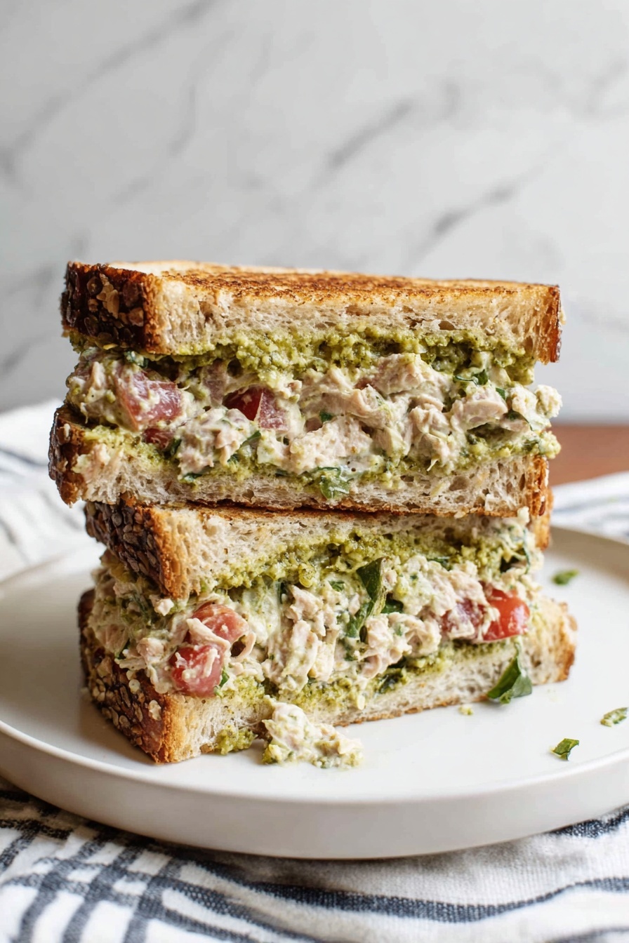 A sandwich cut in half and stacked on a white plate, showing three layers: the top and bottom are toasted multigrain bread with visible seeds and a golden-brown crust; the first filling layer is a thick, creamy tuna salad mixed with small pieces of tomato and green herbs, and the second filling layer is a green, chunky spread, likely pesto, spread on the bread. The sandwich looks moist and slightly messy with some filling falling onto the plate. The background has a white marbled texture and a striped cloth is partly visible behind the plate. Photo taken with an iphone --ar 2:3 --v 7