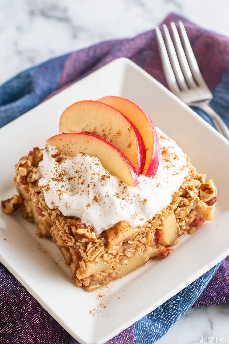 The dish shows one thick square layer of baked apple oatmeal with visible chunks of apple and walnuts. On top, there is a thick white swirl of whipped cream sprinkled with cinnamon powder, and several thin slices of red apple fanned out on one side. The dish is served on a white square plate, placed on a white marbled texture surface, with a silver fork and stacked blue and purple napkins in the background. Photo taken with an iphone --ar 2:3 --v 7