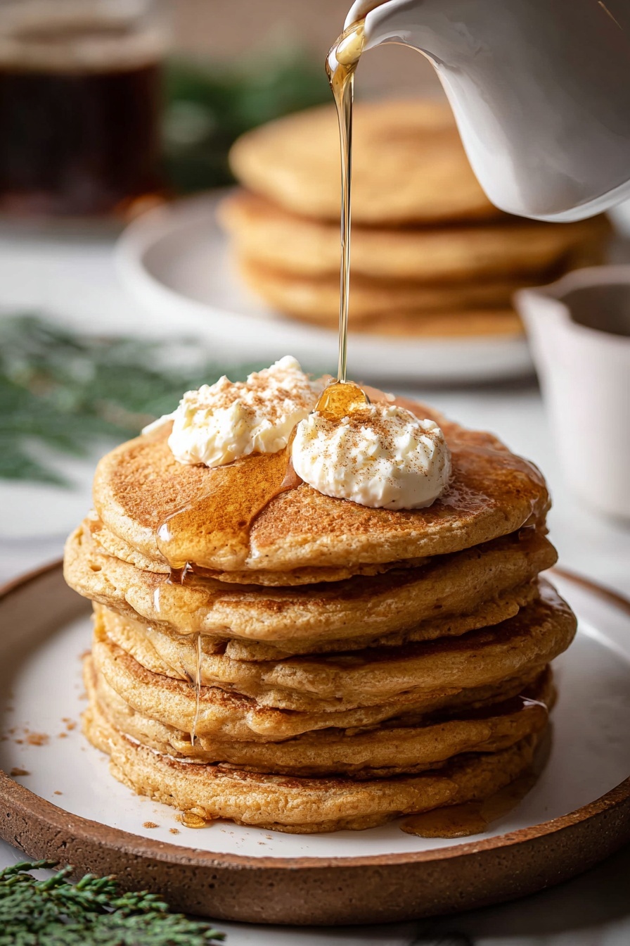 A stack of six thick, golden brown pancakes sits on a white plate with a visible fluffy texture. On top, there are two small dollops of creamy butter lightly sprinkled with cinnamon. A stream of amber syrup is being poured from a white jug, slowly flowing over the butter and down the sides of the stack. In the background, there is a blurry stack of more pancakes on a white plate and a glass with a dark drink. The whole scene rests on a white marbled surface with some green herbs adding a soft touch of color in the background. photo taken with an iphone --ar 2:3 --v 7