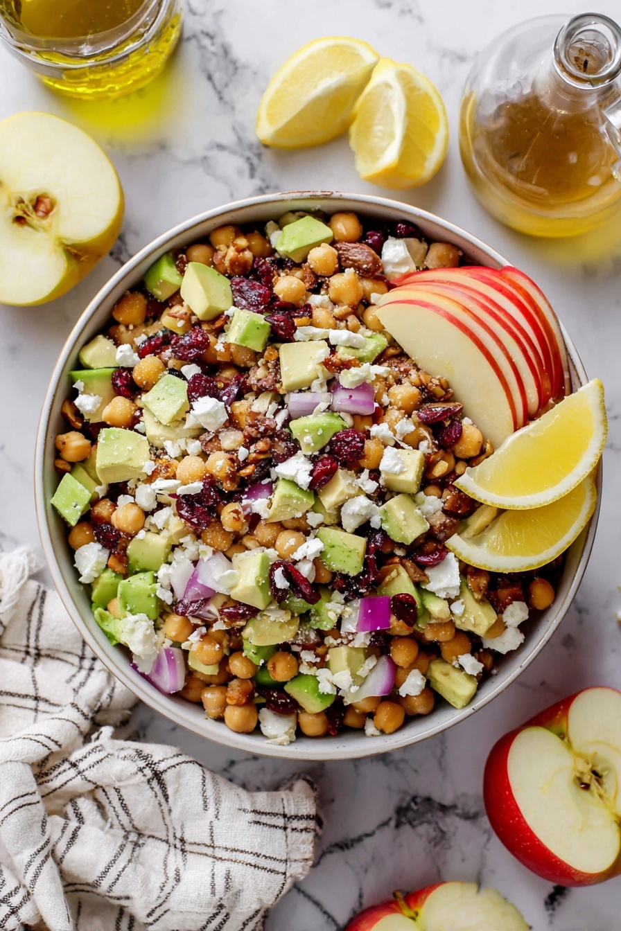 A white bowl filled with a colorful salad showing many layers and textures: the bottom layer is light beige chickpeas, mixed with bright green avocado chunks and red apple pieces, topped with small pieces of white cheese and scattered dark red dried cranberries. There are also small bits of purple onion and broken pecans spread over the salad. On the left side of the bowl, there are three lemon wedges placed on top of the salad. The bowl sits on a white marble surface, surrounded by a glass bottle of olive oil on the top left, half a red and yellow apple on a wooden board at the top right, and a sliced lemon below the bowl. A white and gray striped cloth is at the bottom left corner of the image. photo taken with an iphone --ar 2:3 --v 7