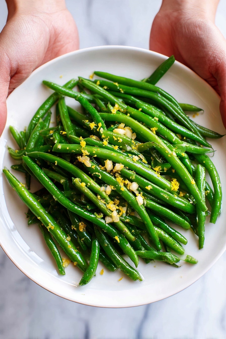 A white plate is filled with bright green cooked green beans piled in one layer, with small bits of chopped garlic and tiny yellow lemon zest scattered evenly on top. Two woman's hands hold the plate from either side against a white marbled surface. In the blurry background, there is a small white bowl holding two lemon wedges. The green beans look shiny and fresh with a simple, clean presentation. photo taken with an iphone --ar 2:3 --v 7