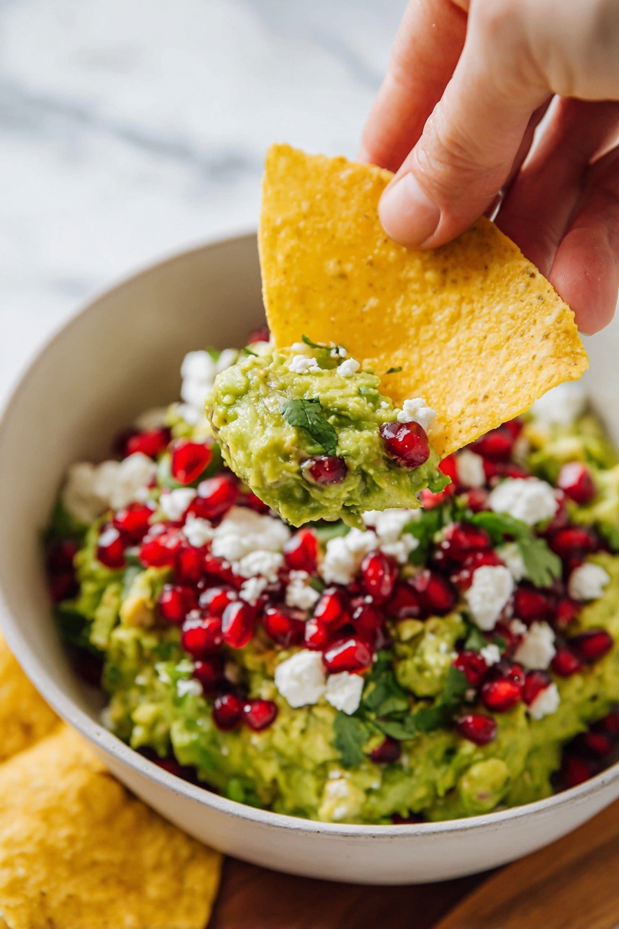 A white bowl filled with green guacamole spread evenly inside, topped with bright red pomegranate seeds and small white cheese crumbles, garnished with a few fresh green cilantro leaves. A large yellow tortilla chip is dipped into the guacamole on one side of the bowl. The bowl sits on a round wooden board surrounded by several yellow tortilla chips spread around the edge. Some fresh cilantro leaves lie next to the bowl on the board. The background is a white marbled texture with a dark blue cloth peeking from the left side. Photo taken with an iphone --ar 2:3 --v 7