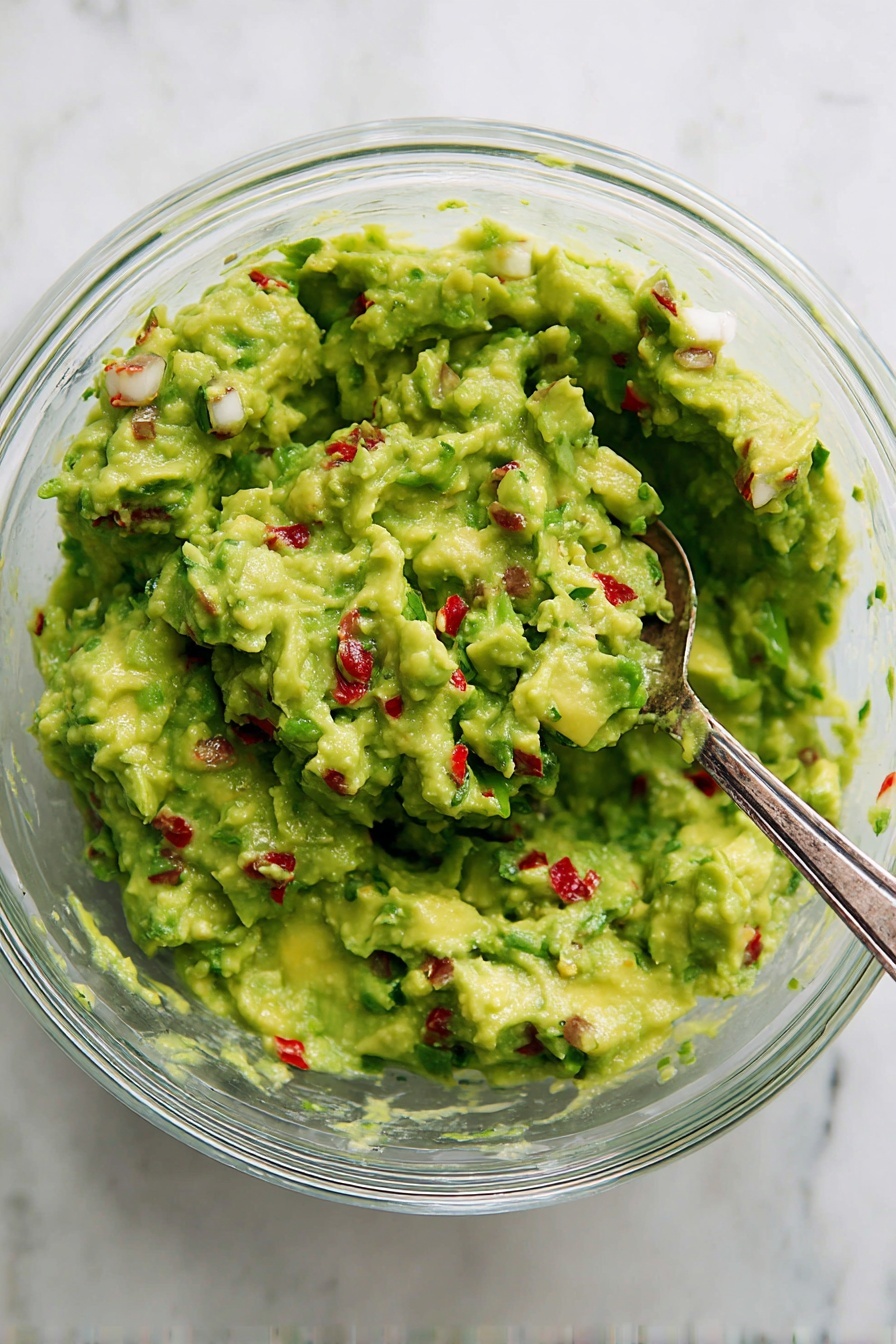 A clear glass bowl filled with chunky green guacamole that shows mix of smooth and small pieces texture, scattered with tiny red bits and some white pieces, a silver spoon is partially dipped on the right side of the bowl, all placed on a white marbled surface photo taken with an iphone --ar 2:3 --v 7