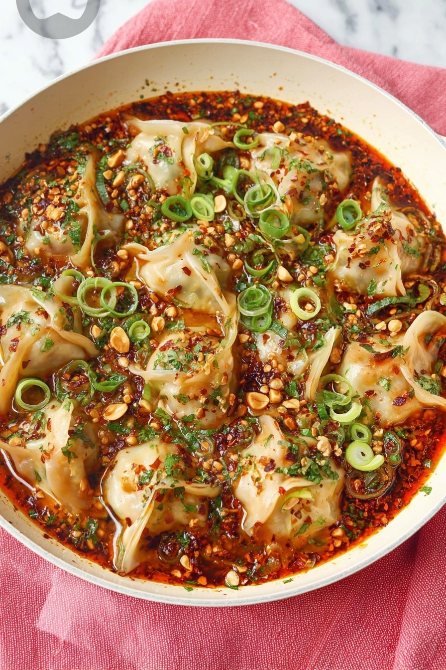 A white deep pan is filled with several dumplings layered on a thick red-orange sauce that looks spicy and oily. The dumplings have light golden edges with soft folds and are topped with bright green chopped scallions and cilantro leaves scattered around. There are small bits of toasted nuts and chili flakes spread over the dumplings and sauce. A woman's hand holding a gold spoon is lifting one dumpling from the pan. The pan is placed on a soft pink cloth on a white marbled surface, with part of a white plate with green dots and a pair of wooden chopsticks visible near the bottom. In the background, a green textured mug is partially visible. photo taken with an iphone --ar 2:3 --v 7