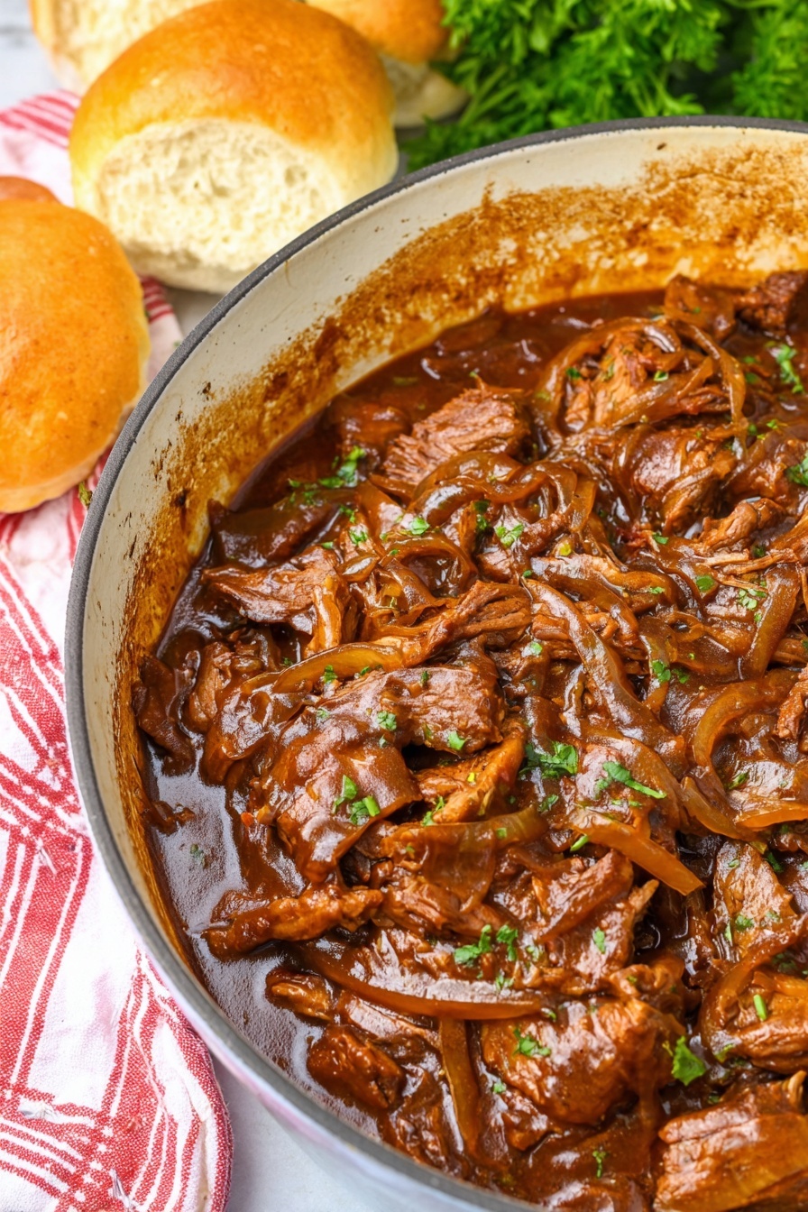 A close-up view of a white enamel pot filled with several layers of thin, brown cooked meat pieces mixed with caramelized onions and covered in a thick, shiny brown sauce. The edges of the pot show the sauce stains, adding texture to the sides. To the upper left, three soft round golden brown bread rolls rest next to the pot on a red and white striped cloth, all set on a white marbled surface. Fresh green parsley is partly visible in the top right corner. Photo taken with an iphone --ar 2:3 --v 7