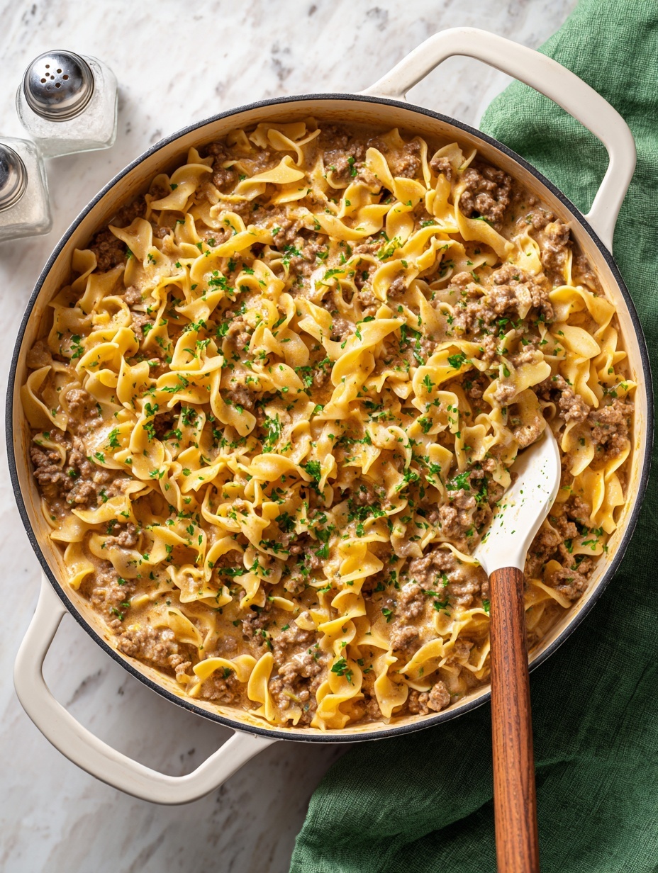 A close top view of a white pan filled with creamy pasta mixed with browned ground meat and small chopped green herbs scattered on top, with a white spatula with a wooden handle resting inside the pan on a white marbled surface; the pasta is light yellow with a soft texture, and the meat is crumbly and brown, blending into the creamy light beige sauce, photo taken with an iphone --ar 2:3 --v 7