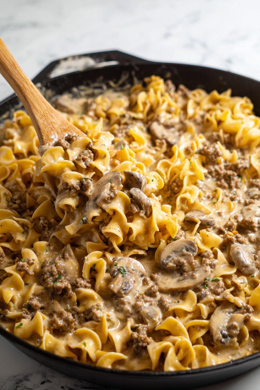A skillet filled with a creamy noodle dish shows three main layers: the top layer is yellowish egg noodles with soft, round edges, mixed gently with small pieces of brown ground meat and beige, thinly sliced mushrooms. The creamy sauce, light tan in color, coats every piece giving a smooth and slightly shiny texture. A wooden spoon lifting a scoop in the center highlights the noodles, meat, and mushrooms closely clumped together with the sauce. The skillet is black with curved edges, set on a surface with a white marbled texture. Photo taken with an iphone --ar 2:3 --v 7