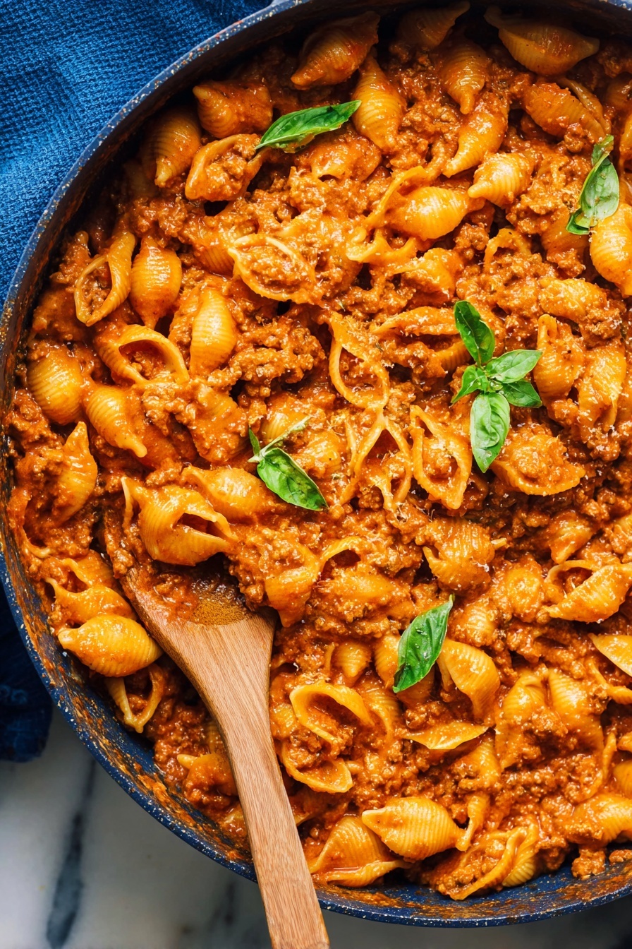 A close-up view of a large pan filled with shell pasta mixed with thick orange-red meat sauce, evenly coating each pasta piece. The sauce has a slightly chunky texture from ground meat, creating a rich, hearty look. Scattered on top are a few fresh, bright green basil leaves, adding a fresh contrast to the warm colors. A wooden spoon is partially submerged in the pasta on the left side of the pan. The pan sits on a white marbled surface, and a blue cloth is visible on the upper left edge of the frame. Photo taken with an iphone --ar 2:3 --v 7