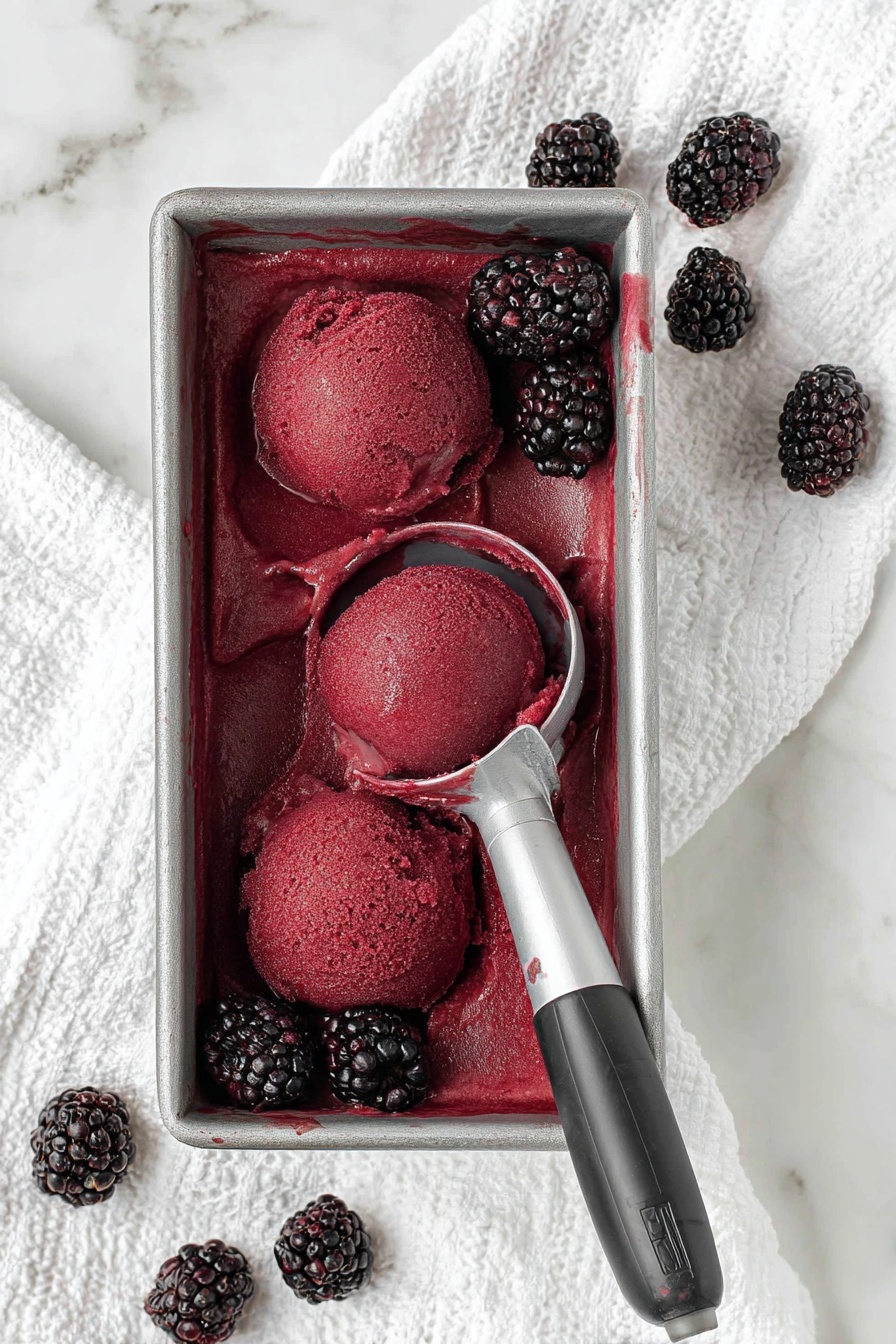 A rectangular metal container holds a deep red-purple blackcurrant sorbet with a smooth, slightly icy texture. Three rounded scoops are carved into the sorbet, with one scoop resting on a silver scooper with a black handle placed on the right side of the container. Around the sorbet, several fresh blackberries with a glossy, bumpy surface are placed both inside and outside the container on a white textured cloth. The background is a white marbled surface. photo taken with an iphone --ar 2:3 --v 7