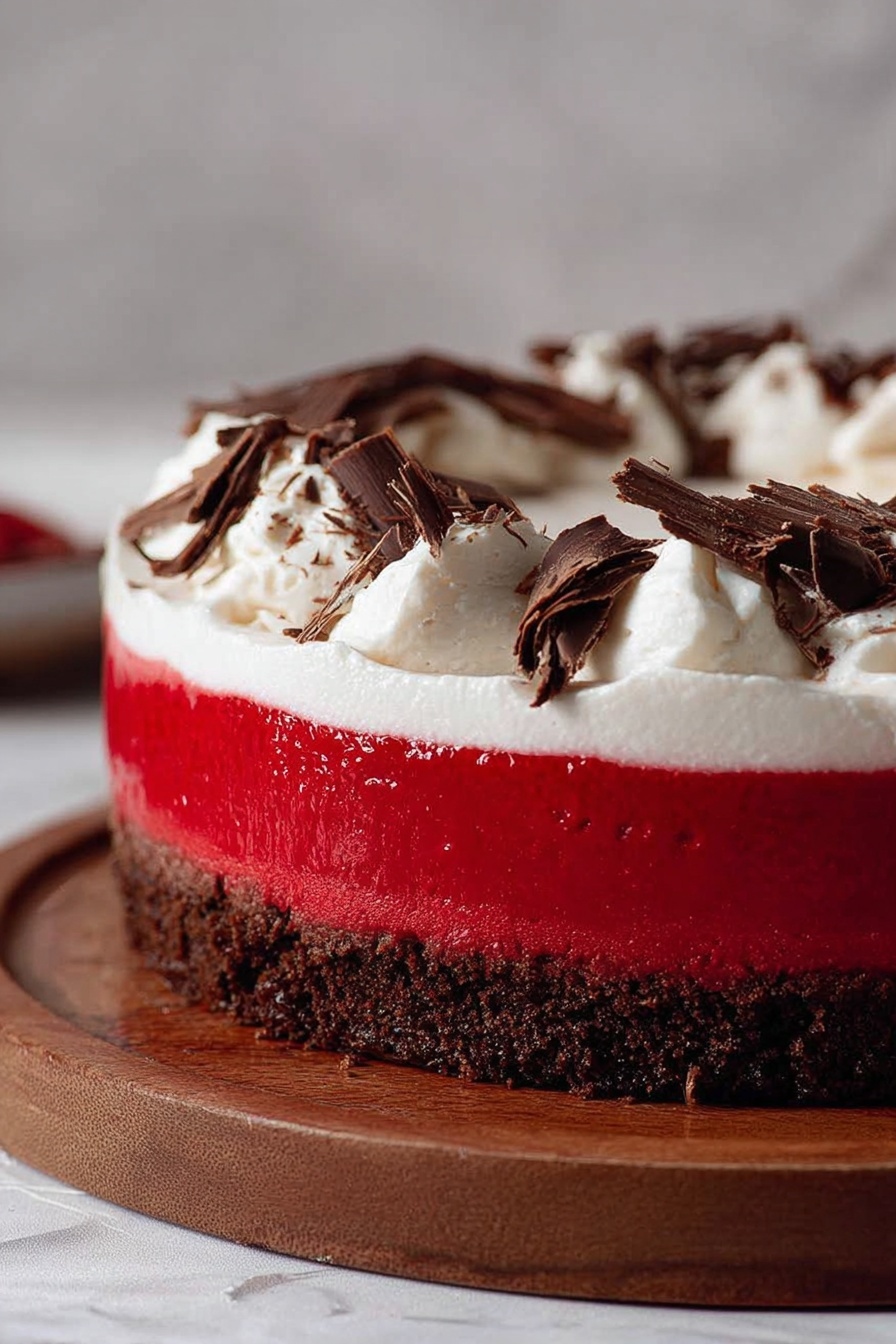 The image shows a three-layer dessert on a round wooden board. The bottom layer is a dark brown, crumbly crust. The middle layer is thick, smooth, and bright red with a shiny surface. The top layer is a thick swirl of white whipped cream, topped with thin dark brown chocolate shavings scattered over it. The background is a soft blur with a white marbled texture surface underneath. photo taken with an iphone --ar 2:3 --v 7