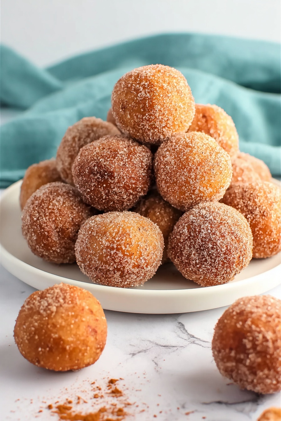 The image shows a white plate piled with many round doughnut balls coated evenly in cinnamon sugar. The doughnut balls have a golden-brown color with a slightly rough texture from the sugar coating. Some doughnut balls are scattered on the white marbled surface near the plate, with a few pieces of sugar and cinnamon around them. The scene also includes part of a blue cloth to the left side. The lighting highlights the warm tones and texture of the doughnut balls, giving them a fresh and inviting look. photo taken with an iphone --ar 2:3 --v 7
