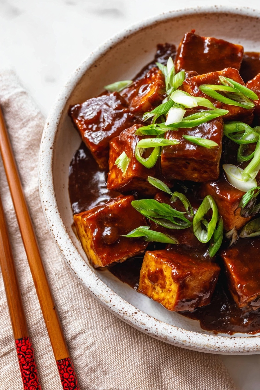 The image shows a close-up of square pieces of tofu coated in a thick, shiny dark brown sauce, filling most of a shallow white speckled bowl. Bright green sliced scallions are scattered on top, adding a fresh touch. The bowl rests on a soft beige cloth with a pair of wooden chopsticks featuring red decorations beside it, all set on a white marbled surface. The tofu pieces have a smooth yet slightly textured look with sauce pooling slightly beneath them. photo taken with an iphone --ar 2:3 --v 7
