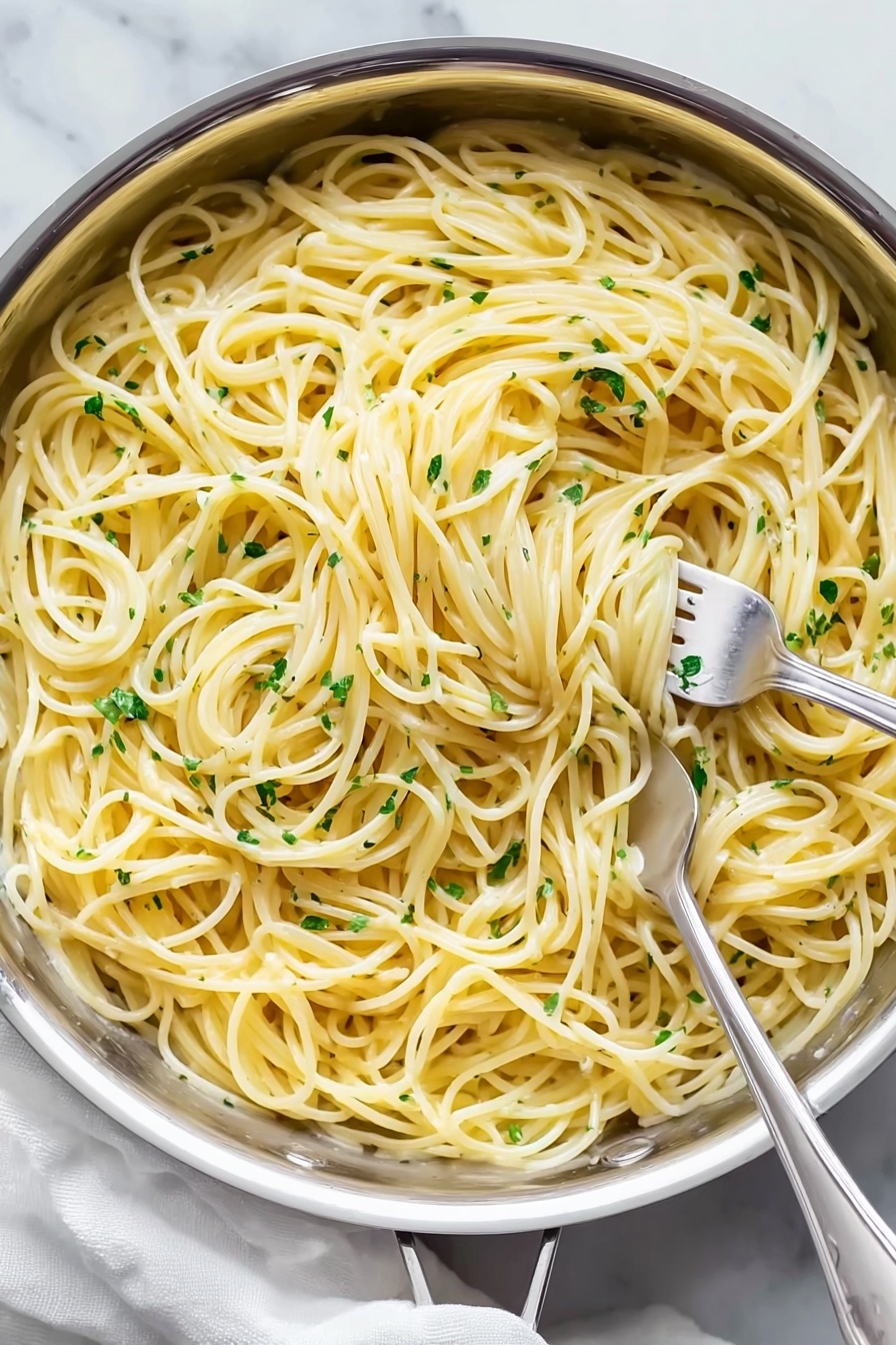 A large silver pan filled with creamy spaghetti pasta swirled into loose nests with two silver forks twirling the noodles. The pasta strands are pale yellow and smooth, mixed evenly with small bits of green herbs scattered throughout. The pan sits on a white marbled surface with a soft white cloth partially visible underneath. The noodles appear soft and coated with a light sauce that gives a slight shine. Photo taken with an iphone --ar 2:3 --v 7